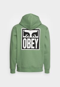 Grön hoodies i mjukt tyg. Har en stor svart och vit "OBEY" grafik på ryggen med handdesigns. Modellen har en huva och ribbade ärmslut.