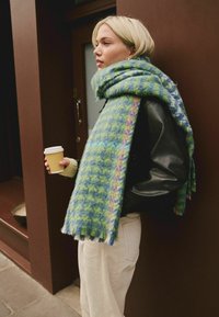 Lys hår person i sort læderjakke og oversized grøn-blå ternet tørklæde holder en takeaway kaffe kop, mens de står ved en brun væg.