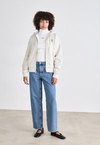 Lichtgrijze rits hoodie met trekkoorden, gedragen over een witte coltrui, gecombineerd met blauwe high-waisted jeans en zwarte flats.