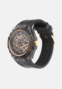 Selge kell läbipaistva korpuse, musta kummirihma, kuldsete detailide ja skeleton-dial'iga, mis toob esile keerukaid mehhaanilisi üksikasju.