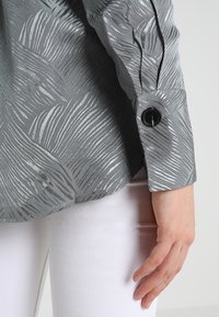 Graue Bluse mit metallic Wellenmuster, ausgestattet mit einem schwarzen Knopf am Umschlag, kombiniert mit weißen Hosen. Textiles Material.