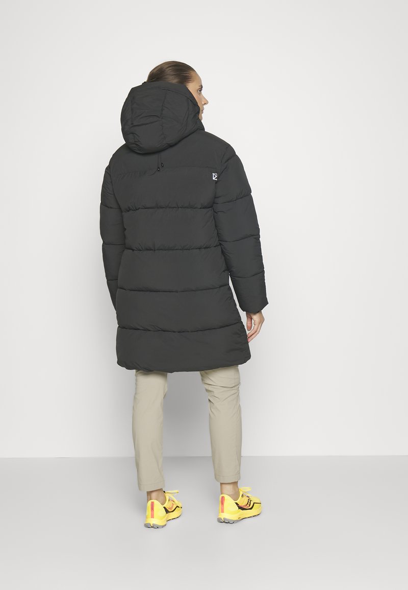 Schwarze Pufferjacke mit Kapuze und horizontaler Steppung. Kombiniert mit beigen Hosen und leuchtend gelben Sneakers; minimale Markenkennzeichnung.