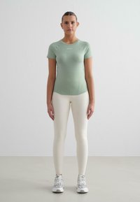 Mintgrön figurnära kortärmad topp med slät textur, kombinerad med krämfärgade leggings. Modellen bär vita träningsskor och står mot en ljus bakgrund.