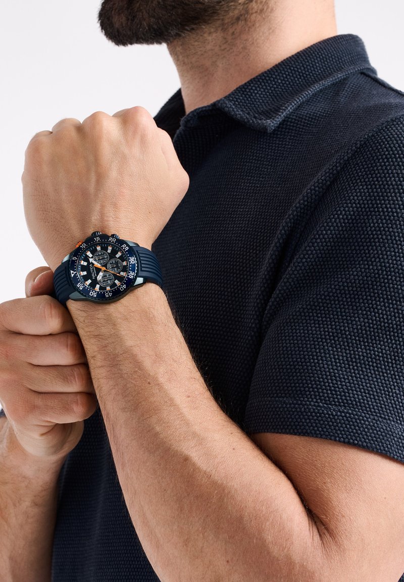 Jūras spēku gumijas pulksteņi ar strukturētu siksniņu, liela apaļa virsma, melns ciparnīca un oranžas akcenti. Valkāt uz rokas virs tumšas polo krekla.