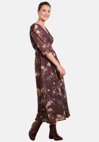 Langes, fließendes Kleid in dunklem Mauve mit floralem Muster, ausgestattet mit einem V-Ausschnitt und Puffärmeln, kombiniert mit braunen Knöchelstiefeln.