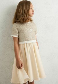 Un vestido top de lentejuelas beige con una falda plisada color crema, que presenta mangas cortas y una textura suave. El dobladillo es ligeramente acampanado.