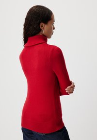 Maglione a collo alto rosso realizzato in tessuto morbido, caratterizzato da orlo e polsini a costine, forma aderente e colletto alto, indossato con jeans in denim scuri.