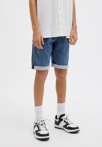 Shorts di jeans blu scuro con risvolti, abbinati a calzini bianchi e sneaker in bianco e nero. Camicia bianca a maniche corte e con texture.
