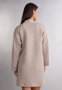 Světle béžové šaty s texturovaným zigzag vzorem, dlouhými rukávy a kulatým výstřihem. Měkká látka sahá těsně nad kolena.