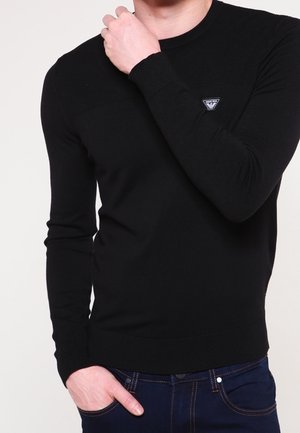 Man met een zwarte, lange mouwen trui met een klein logo op de borst, de ene hand past de kraag aan, de andere in de zak van een blauwe spijkerbroek.