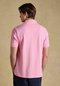 Jeune homme aux cheveux bruns courts portant un polo à manches courtes rose clair et un pantalon foncé, vu de dos sur un fond beige.