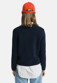 Marineblauer Pullover mit strukturierter Strickoptik, über einem weißen Hemd mit Streifenmuster getragen, kombiniert mit hellblauen Jeans und einer orangefarbenen Mütze.