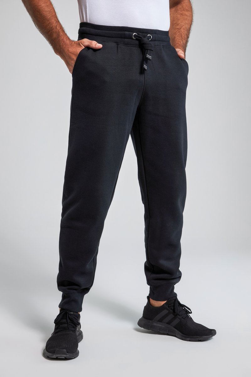 Uomo che indossa pantaloni da jogging neri con coulisse in vita e sneakers nere, in piedi con le mani nelle tasche contro uno sfondo semplice.