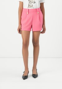 Shorts taillés roses à taille haute, avec deux boutons dorés et des poches latérales, associés à un t-shirt graphique et des talons noirs.