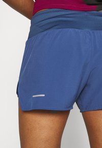 Close-up van een persoon die blauwe sportshorts draagt met een donkere tailleband en een zichtbare reflecterende strip aan de linkerkant.
