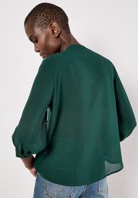 Blusa verde di chiffon con un design rilassato, maniche arricciate e un sottile dettaglio di schiena aperta. Abbinata a jeans in denim blu chiaro.