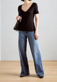 Top nero a maniche corte con bordo in pizzo su un colletto a V, abbinato a jeans a gamba larga blu chiaro. La modella tiene una piccola borsa nera.