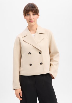 Kobieta o jasnobrązowych włosach, ubrana w beżową dwurzędową kurtkę i czarne spodnie, stojąca na jednolitym białym tle.