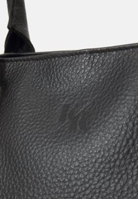 Μαύρη δερμάτινη τσάντα χειρός με υφή στην επιφάνεια, κοντό λουρί και ανάγλυφο λογότυπο. Ο σχεδιασμός είναι μινιμαλιστικός και λειτουργικός.