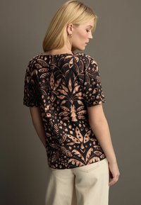 Haut noir à manches courtes avec un motif botanique beige, offrant une coupe décontractée et un ourlet arrière arrondi, fabriqué en tissu texturé léger.