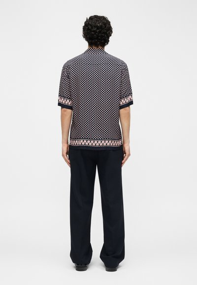 Άνδρας με σγουρά μαλλιά, φοράει κοντομάνικο πουκάμισο με σχέδια από ροζ καρδιές και μαύρο παντελόνι με φαρδιά μπατζάκια, στέκεται γυρισμένος με την πλάτη.