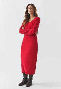 Rode jurk met lange mouwen, een V-hals, een aansluitende silhouette en een geribbelde textuur. Gedragen met kniehoge, bruine leren laarzen.