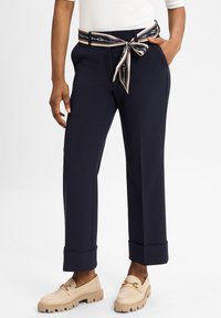 Marineblauwe tailorpantalon met een brede pijpontwerp, voorzien van een gestreepte riem die in de taille is geknoopt en opgerolde boorden. Gemaakt met beige instappers.