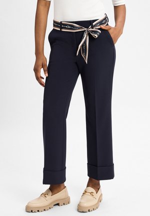 Marineblaue, taillierte Hosen mit weitem Bein, versehen mit einem gemusterten Gürtel, der an der Taille gebunden ist, sowie umgeschlagenen Säumen. Kombiniert mit beigen Slippern.