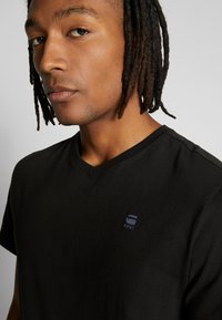 Black v-neck t-shirt made of soft fabric, featuring a small blue logo on the left chest. Simple, fitted design with short sleeves.