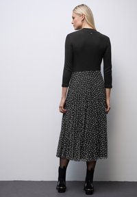 Vrouw met blond haar draagt een zwart shirt met lange mouwen en een zwarte midi rok met plooien en witte abstracte patronen, staande tegen een effen muur.