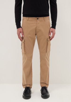 Man draagt beige cargobroek met klepzakken, zwarte schoenen en een zwart shirt met lange mouwen, staand tegen een effen lichte achtergrond.
