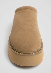 Tan suede slip-on shoe with a rounded toe, textured upper, and a thick, light-colored sole. Features stitching detail around the edges.