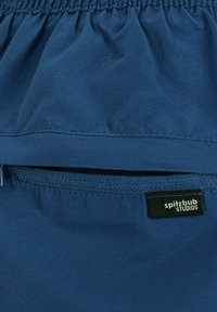 Marinefarbene Stoff mit einer zerknitterten Textur. Verfügt über eine mit Reißverschluss versehenen Tasche mit einem schwarzen Etikett, auf dem "spitzbub STUDIOS" steht. Einfaches, funktionales Design.