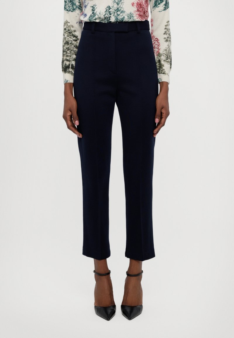 Tailored navy trousers with a smooth texture, slim fit, and ankle length. Styled with black high heels and a patterned top.