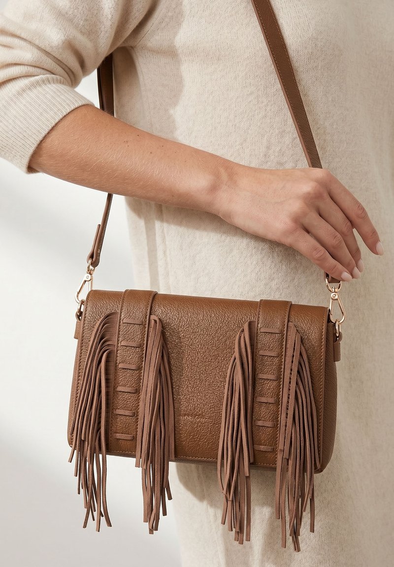 Sac bandoulière en cuir marron avec des détails à longues franges, tenu par une personne portant un haut beige à manches longues et une robe.