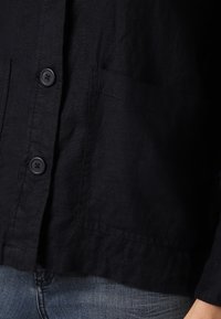 Svart linne skjorta med avslappnad passform, utrustad med två knappar, en bröstficka och subtil textur. Kombinerad med blå denimjeans.