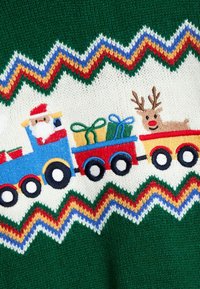 Grüner gestrickter Pullover mit einem weißen Zickzackmuster. Ein blauer Zug mit dem Weihnachtsmann, ein roter Zug mit Rentieren und bunte Geschenke sind gestickt.