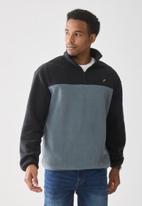 Sudadera de forro polar en azul claro y negro. Cuello con cremallera, mangas largas, tejido texturizado, con un pequeño logo dorado en el pecho. Ajuste casual.