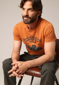 Oranje katoenen t-shirt met grafische print van een vintage vrachtwagen, gecombineerd met donkergrijze broek, zittend op een bruine leren stoel.