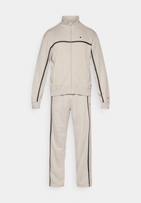 Μπεζ φόρμα κατασκευασμένη από μαλακό ύφασμα. Το μπουφάν διαθέτει ψηλό γιακά και κλείσιμο με φερμουάρ, με μαύρες ρίγες κατά μήκος των μανικιών και των παντελονιών.