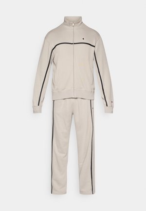 Béžová súprava z jemnej tkaniny. Bunda má vysoký golier a zapínanie na zips, s čiernymi pásikmi pozdĺž rukávov a nohavíc.