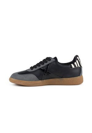 Zapatilla de perfil bajo en negro y gris con suela de goma marrón, cordones negros y detalle en la lengüeta del talón a rayas en negro y blanco.