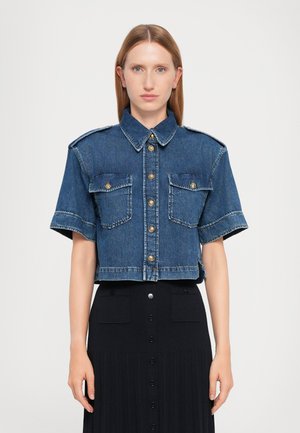Lyhyt denim-paita keskisinisessä värissä, jossa on kaulus, lyhyet hihat, kultaiset nappikiinnitykset ja kaksinkertaiset povitaskut.