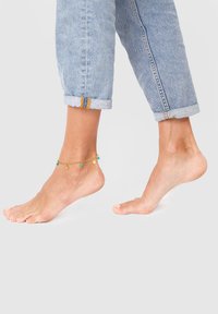 Bracciale d'oro alla caviglia con perline verdi e blu, indossato su un piede nudo. I jeans sono risvoltati alla caviglia, rivelando i dettagli del bracciale.