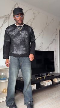 Homme portant un pull noir en maille, un jean bleu, une casquette noire et des lunettes se tient dans le salon avec une télévision et une bibliothèque.