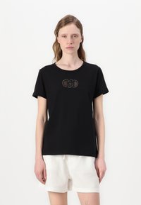 Maglietta nera in cotone a maniche corte, con scollo rotondo e un design circolare con dettagli di perline sul petto. Abbinata a pantaloni corti bianchi.