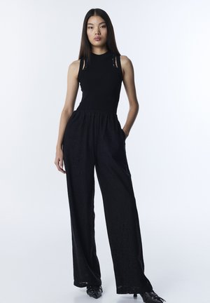 Kobieta stojąca w czarnym, bezrękawnikowym topie i szerokich, czarnych spodniach z koronki z rękami w kieszeniach na białym tle.