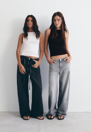 Dve ženski stojita ob beli steni, oblečeni v brezrokavnike (belo in črno), široke kavbojke z razširjenimi hlačnicami in črne japonke, z rokami v žepih ali v zanki pasu.