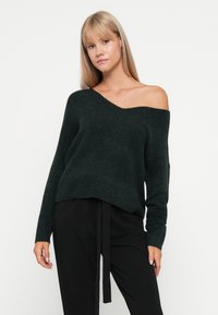 Donkergroene off-shoulder sweater van zacht breimateriaal, met een relaxte pasvorm en geribbelde manchetten. Gescumpleerd met zwarte broek.