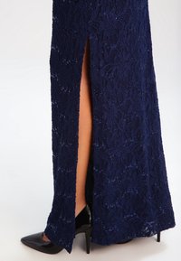 Marinblå spetsklänning med ett högslitsat design, intrikat blommönster och paljetter; matchad med en svart spetstång högklackad sko.